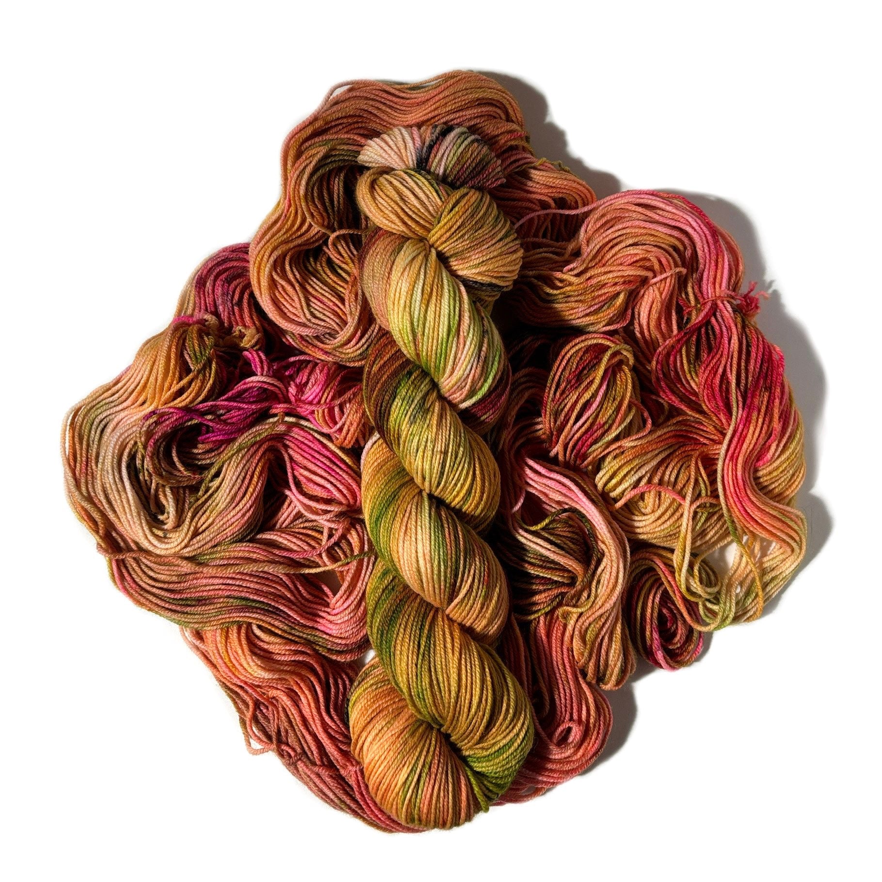 A pile of hand-dyed yarn skeins in a variegated earthy colorway of olive green, terracotta, rust orange, and deep pink, set against a plain white background.