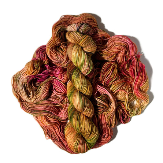 A pile of hand-dyed yarn skeins in a variegated earthy colorway of olive green, terracotta, rust orange, and deep pink, set against a plain white background.
